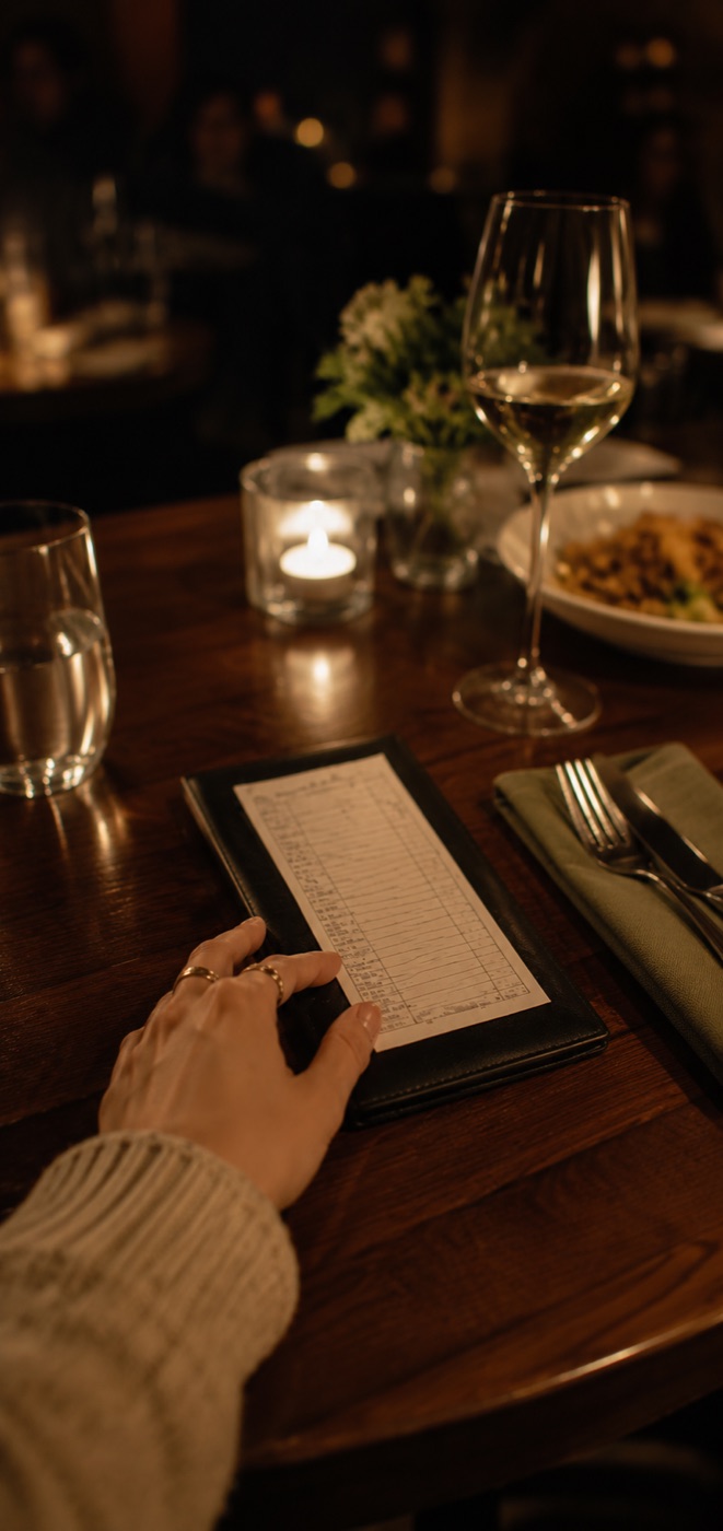 A hand holding a receipt at a candlelit restaurant table with a wine glass and a plate of food in the background.